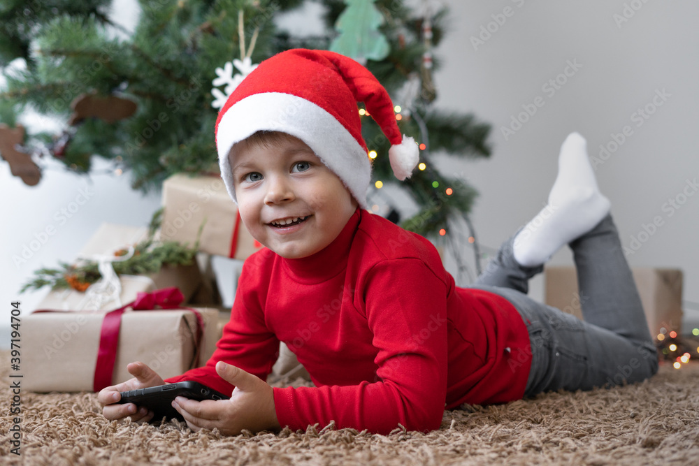 Caucasian boy wearing a red santa claus hat. lies on the carpet holding a box with a gift.