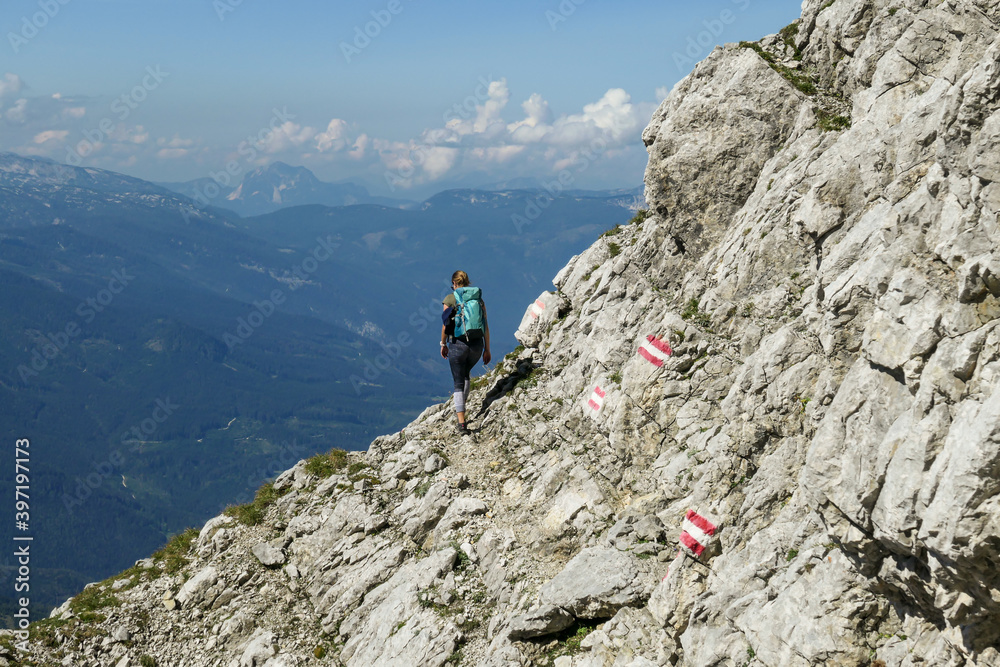 Woman walking on a very narrow and steep pathway along a mountain wall in Austrian Alp, leading to the top of Grimming. The path is marked with red-white-red sign. Dangerous trail. Following the path