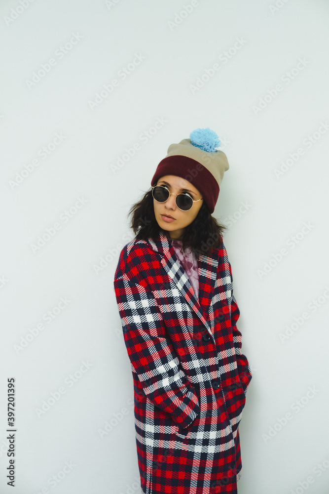 Beautiful fashion model in trendy urban street outfit. Stylish posing. Sunglasses, red coat and funny beanie on white wall background