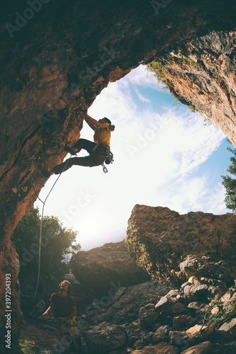The girl climbs the rock in the shape of an arch, A man is belaying a climbing partner.