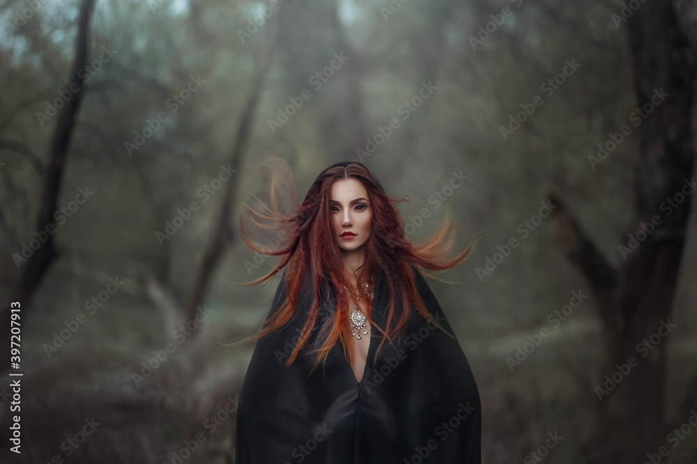 Fototapeta premium Mysterious fantasy gothic woman dark witch obsessed by evil. Red-haired Girl demon in black dress cape hood. Red hair flutters in wind. Dark dense deep forest background, trees. Scleral lenses on eyes
