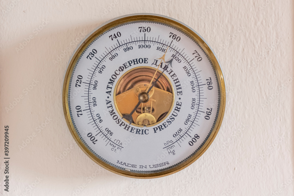 A golden framed, USSR made, atmospheric pressure barometer hanging on a wall. The instrument is inside the glass in white. It shows numbers and has a gold and black arrow A golden framed, USSR made, atmospheric pressure barometer hanging on a wall. The instrument is inside the glass in white. It shows numbers and has a gold and black arrow