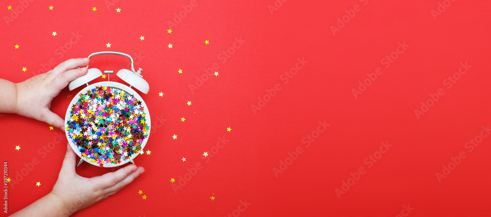 child's hands hold alarm clock with scattered sequins on a red background. christmas, celebration concept. banner