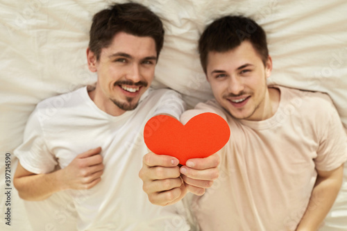 Happy couple of young gay men hide hold red paper heart lying on back out of focus on valentine's day