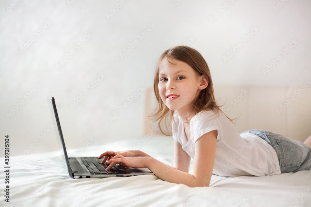 A child girl with a laptop lies on a bed in a real interior in light colors. Distance e-learning at home or online communication with friends and family A child girl with a laptop lies on a bed in a real interior in light colors. Distance e-learning at home or online communication with friends and family