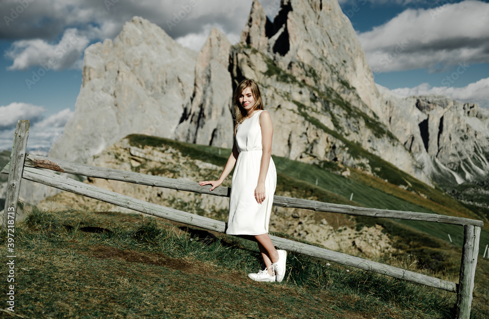 Naklejka premium a young girl in a white dress on the top of the mountain enjoys the sunset against the backdrop of beautiful mountains. travel to the alps. beautiful landscape over the valley. beauty of nature.