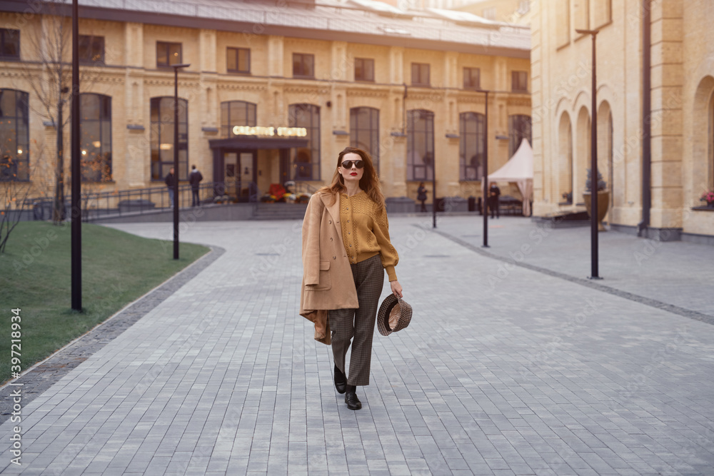 Fashion young woman in sunglasses and hanging on a shoulder autumn beige coat walking on the street happily posing for the camera. Tinted photo. Fashion young woman in sunglasses and hanging on a shoulder autumn beige coat walking on the street happily posing for the camera. Tinted photo.
