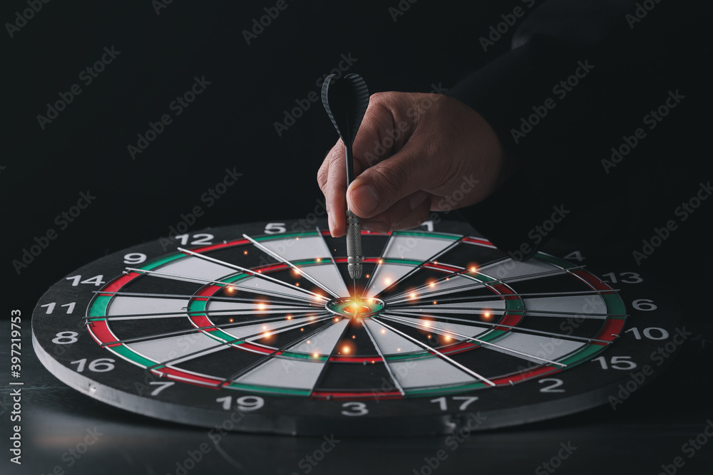 Obraz premium Businessman hand in black suite holding black dart put to center of target board on vintage table meaning of investment and achievement goal. Planning and business strategy concept.
