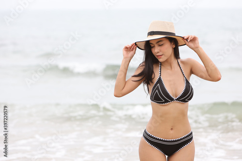 portrait of young asian beautiful and sexy woman in black bikini and white hat with beauty face standing on sandy beach with blue sky and left copy space