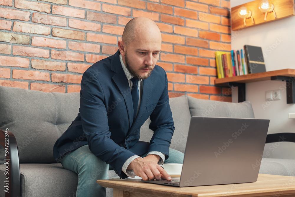 Bearded bald man, businessman or freelancer sitting on a sofa and working on laptop from home, modern interior loft design