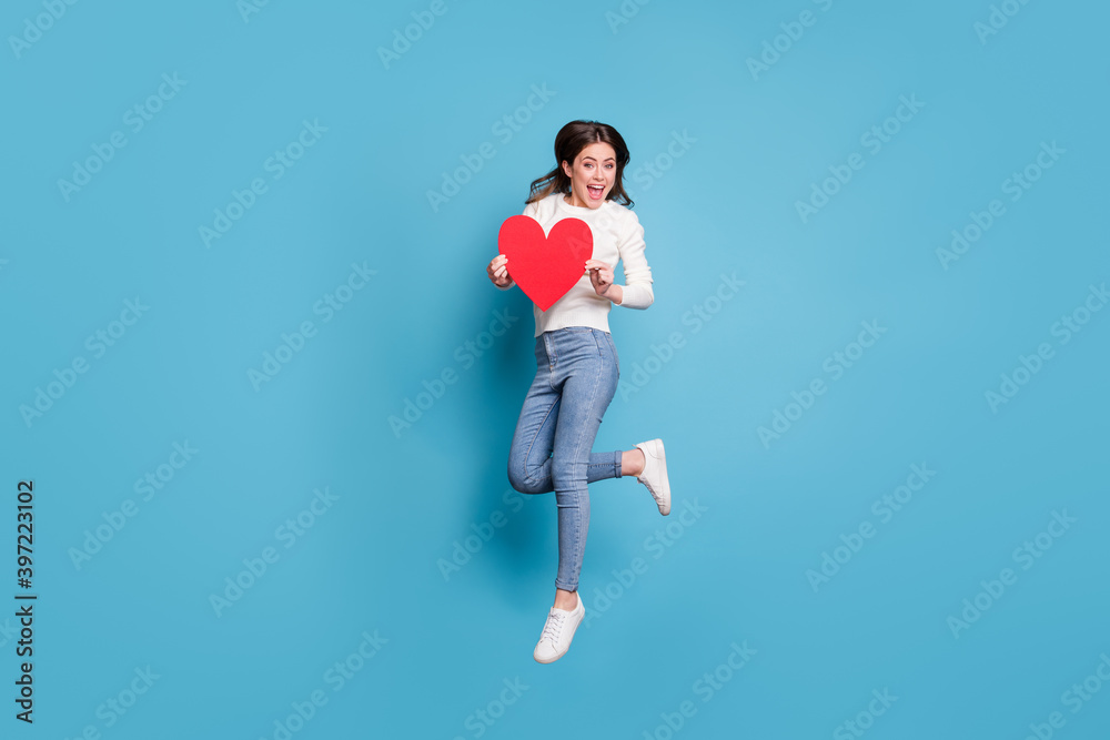 Full length photo of pretty wavy lady wear white sweater jumping holding red paper heart isolated blue color background Full length photo of pretty wavy lady wear white sweater jumping holding red paper heart isolated blue color background