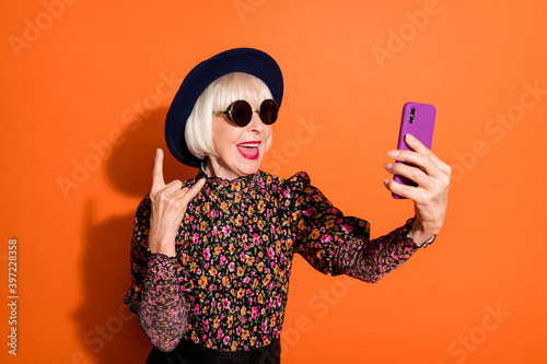 Photo of smiling cheerful happy modern grandmother take selfie on phone show rock n roll sign isolated on orange color background