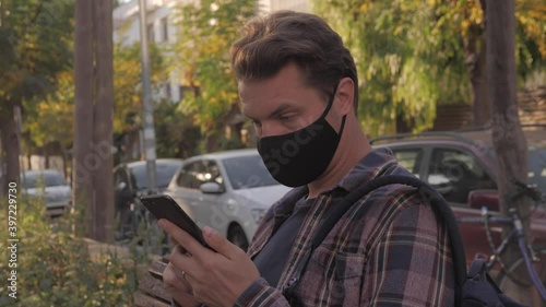 Adult man with surgical medical mask sitting on a bench and using phone, businessman in park relax, safe travel