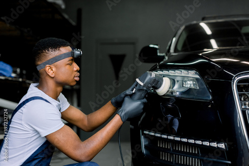 Detailing and polishing of car headlight. Young dark skinned worker in uniform and protective gloves, with orbital polisher in auto service polishing car lights