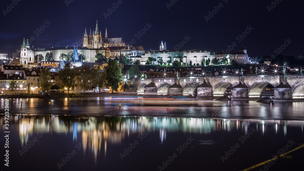 Night cityscape of Prague, Czech Republic. Stone bridge and embankment, blurred reflections in the river, buildings in the night illumination, a huge cathedral in the background