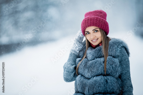 Portrait of a cute girl in a gray fur coat and red hat walking in a winter forest in a snowy day. Winter mood in a pine forest. Christmas time outdoor. 