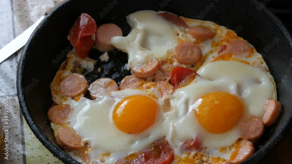 A man eats with a fork a delicious fresh fried egg in a vintage cast-iron frying pan, with seasonings, tomatoes, sweet paprika and sausages, with steam. Morning breakfast in home kitchen. A man eats with a fork a delicious fresh fried egg in a vintage cast-iron frying pan, with seasonings, tomatoes, sweet paprika and sausages, with steam. Morning breakfast in home kitchen.