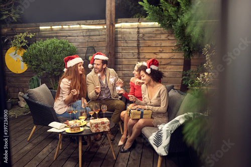 group of young adult multiethnic friends celebrating new year outdoor, sitting in the backyard, having good time, drinking, wine, wearing santa hats with sparkling sticks © luckybusiness