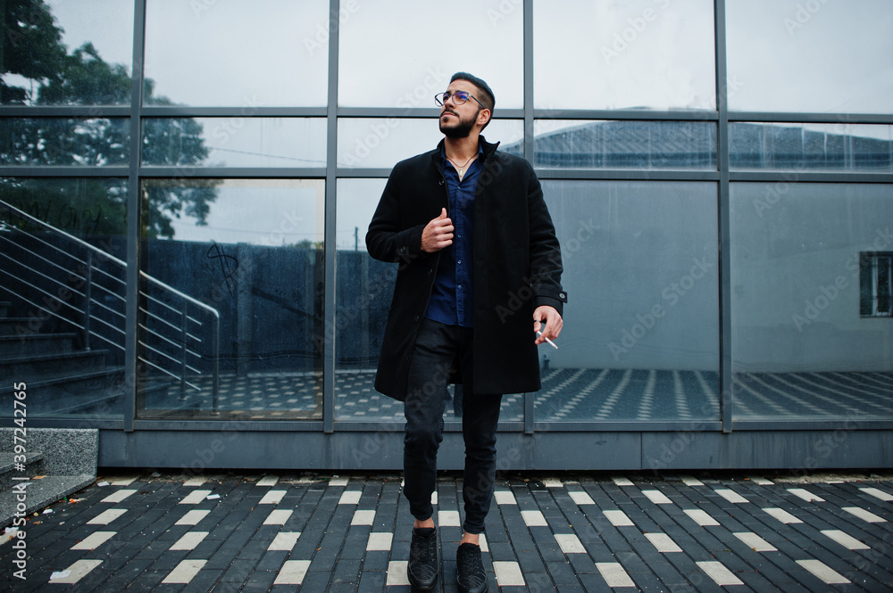 Middle eastern entrepreneur wear black coat and blue shirt, eyeglasses against office building smoking cigarette. Middle eastern entrepreneur wear black coat and blue shirt, eyeglasses against office building smoking cigarette.
