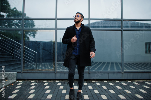 Middle eastern entrepreneur wear black coat and blue shirt, eyeglasses against office building smoking cigarette.