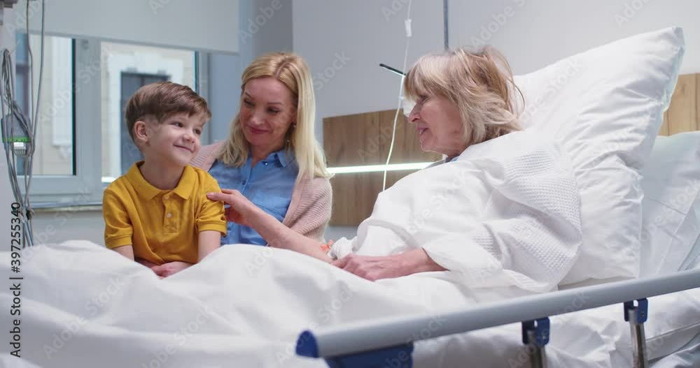 Portrait of happy Caucasian senior woman lying on bed in hospital room and chatting in good mood with little grandson and daughter. Disease treatment. Family relationships. Clinic patient