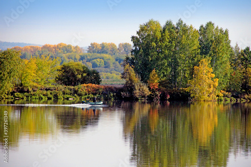 Calmness river surface disturbs driving motor boat. Wonderful landscape of autumn forest on the background.