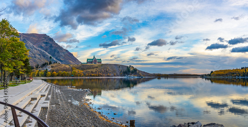 Waterton Lakes National Park lakeshore in autumn foliage season morning. Blue sky, colourful clouds reflect on lake surface like a mirror in sunrise. Fall color landscape. Landmarks in Alberta, Canada