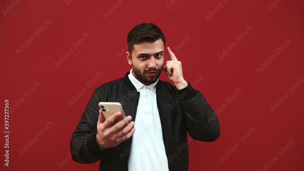 Emotional bearded man uses a smartphone and with a happy face shows a thumb up on a place for advertising on a background of a red wall. Emotional bearded man uses a smartphone and with a happy face shows a thumb up on a place for advertising on a background of a red wall.