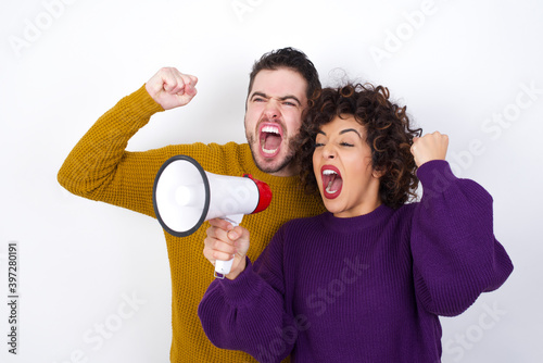 Quadro su tela Young couple wearing knitted sweater standing against white wall communicates shouting loud holding a megaphone, expressing success and positive concept, idea for marketing or sales