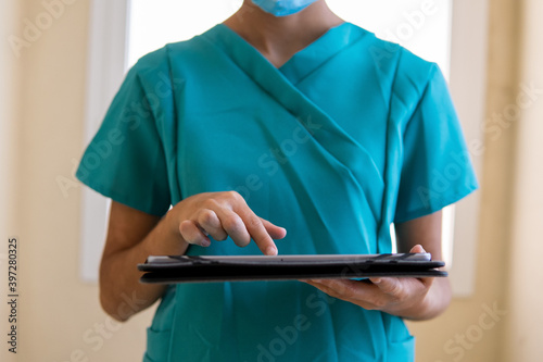 Young nurse in blue scrubs and medical mask reading information on tablet while standing in corridor of modern hospital