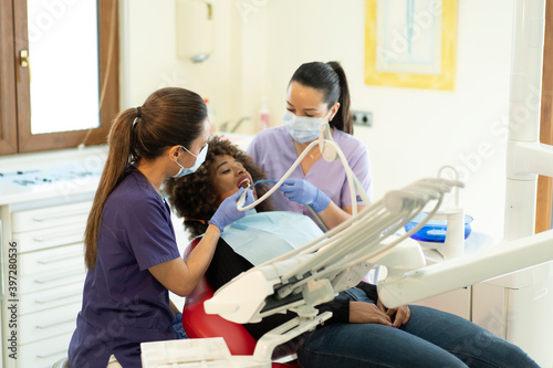 Female assistant and dentist holding instrument to curing teeth of African American lady in clinic