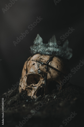 Spooky human skeleton skull with weathered crown placed on pile of ash against black background