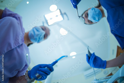 Blurred female dentist with mirror and drill and assistant with suction device standing against clinic ceiling during curing procedure photo