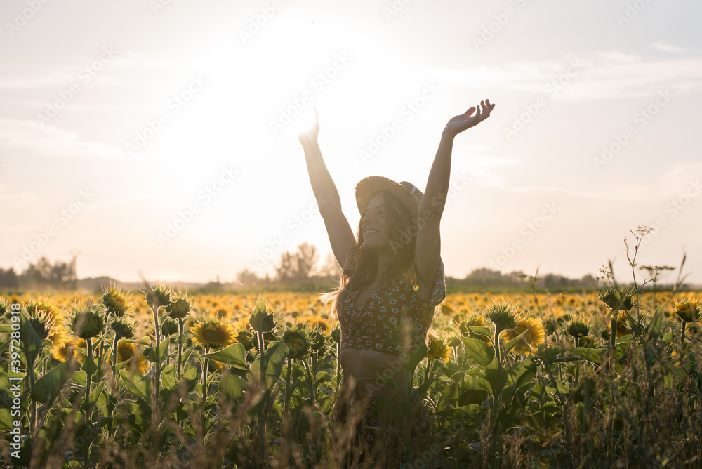 Optimistic young female in summer outfit and hat standing looking away with outstretched arms amidst blooming sunflowers in field and enjoying sunny day Optimistic young female in summer outfit and hat standing looking away with outstretched arms amidst blooming sunflowers in field and enjoying sunny day