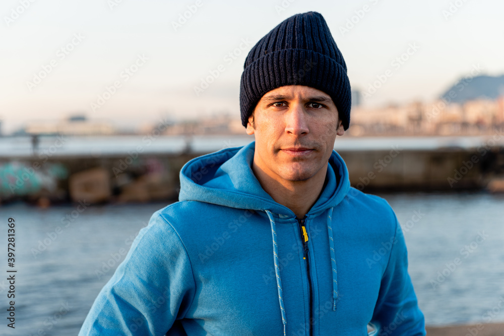 Confident adult ethnic male in warm sportive hoodie and knitted hat looking at camera while standing on embankment near sea at sunset time Confident adult ethnic male in warm sportive hoodie and knitted hat looking at camera while standing on embankment near sea at sunset time