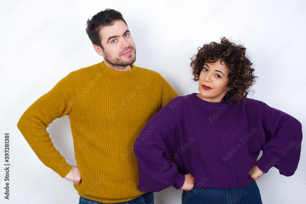 Fototapeta premium Funny frustrated Young couple wearing knitted sweater standing against white wall holding hands on waist and silly looking at awkward situation.