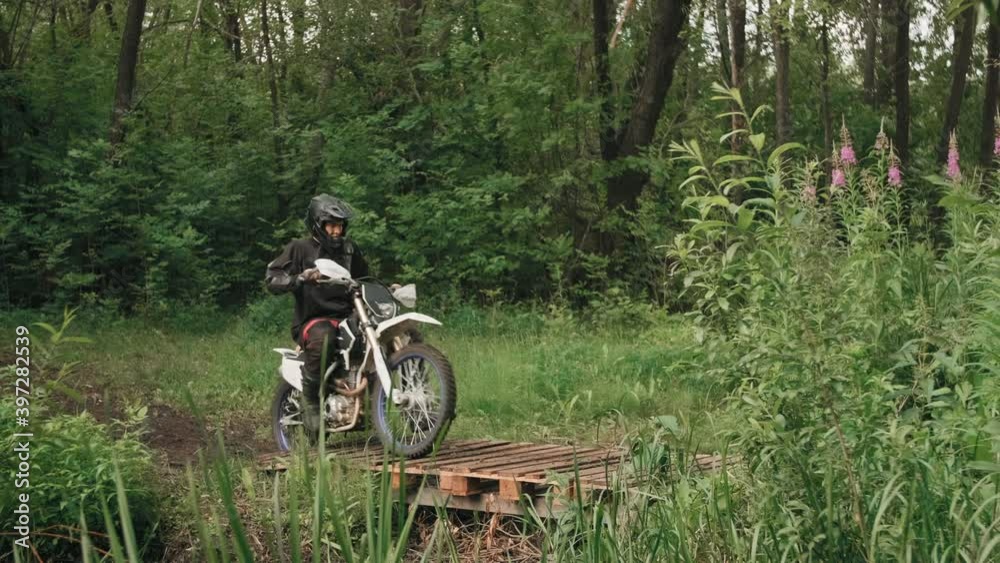 Tracking of men in gear and helmets riding motorcycles through forest, then crossing small wooden bridge Tracking of men in gear and helmets riding motorcycles through forest, then crossing small wooden bridge