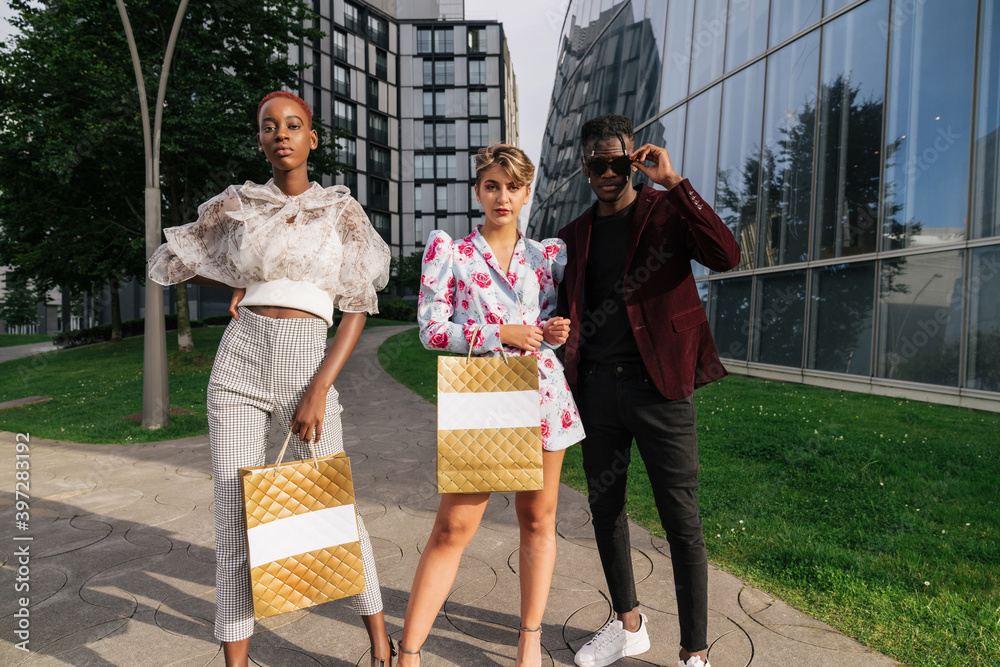 Full length of group of modern millennial multiethnic friends in fashionable outfits holding shopping bags while standing together against contemporary city buildings with glass walls Full length of group of modern millennial multiethnic friends in fashionable outfits holding shopping bags while standing together against contemporary city buildings with glass walls