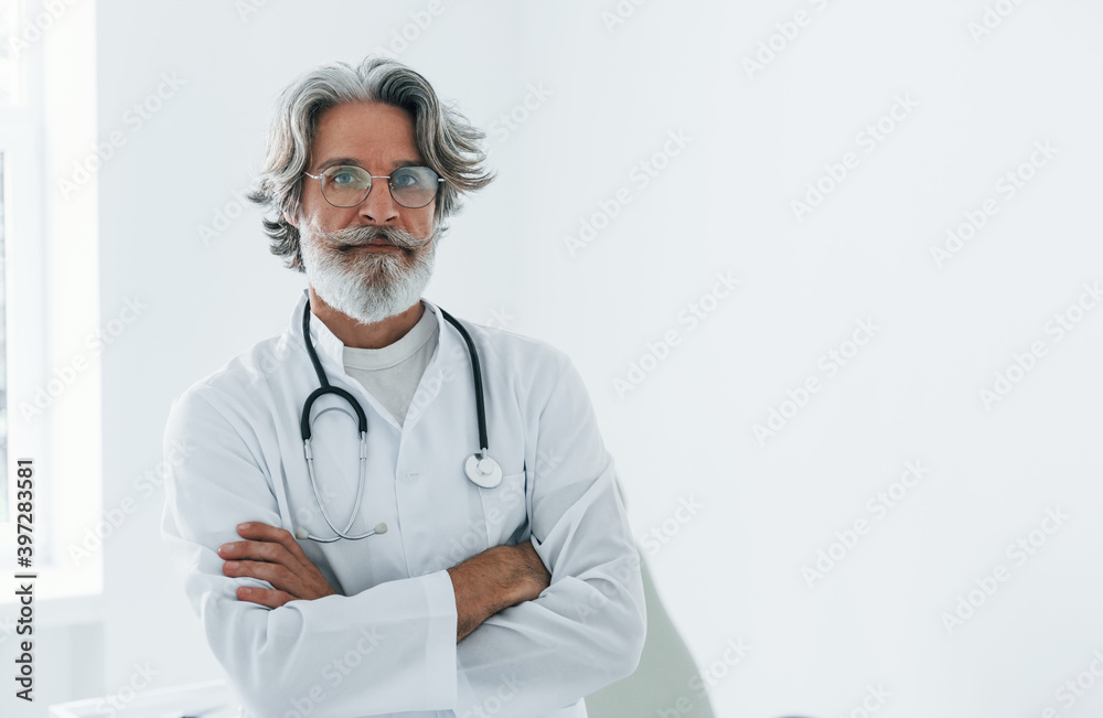 Senior male doctor with grey hair and beard in white coat is indoors in clinic Senior male doctor with grey hair and beard in white coat is indoors in clinic