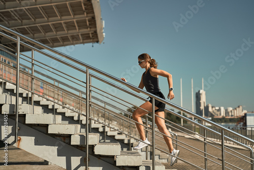 Low angle side view of energetic determined female athlete in sportswear running upstairs on tribune of stadium during outdoor fitness training photo