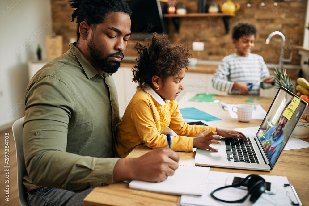 Black working father having video conference and taking notes while babysitting his kids at home. Black working father having video conference and taking notes while babysitting his kids at home.