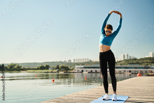 Full body of young fit female in sportswear and sunglasses stretching arms while warming up during fitness workout on wooden pier near river