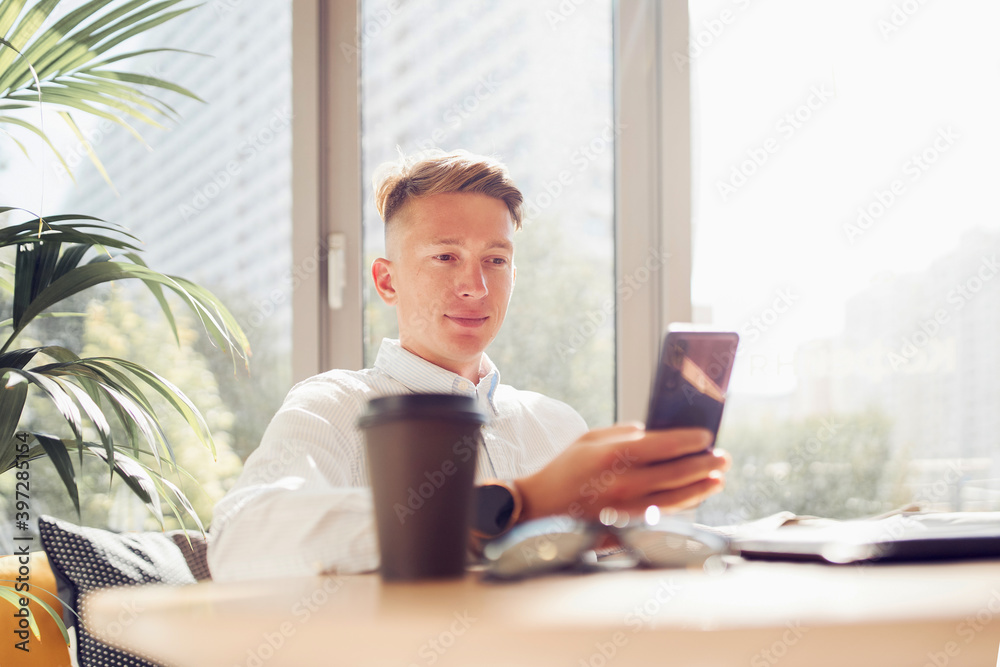 Busy male freelancer sitting at wooden table with takeaway coffee and working on remote project while using smartphone Busy male freelancer sitting at wooden table with takeaway coffee and working on remote project while using smartphone