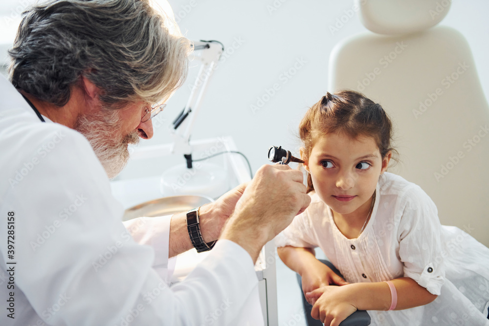 Little girl have a visit. Senior otorhinolaryngologist with grey hair and beard in white coat is indoors in clinic Little girl have a visit. Senior otorhinolaryngologist with grey hair and beard in white coat is indoors in clinic