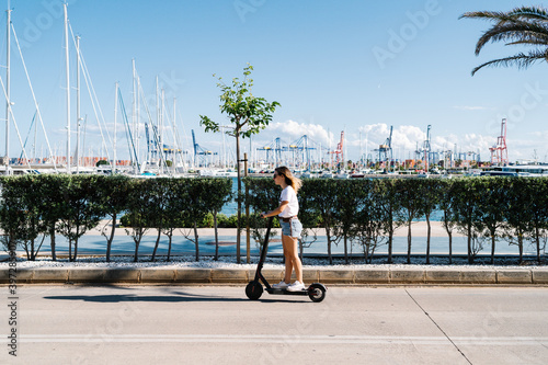 Full body side view of young female in casual outfit riding electric scooter on paved road near urban embankment in sunny summer day photo