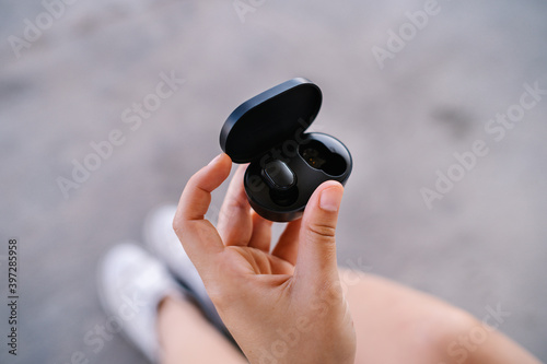 From above crop unrecognizable female holding power bank case with true wireless earbuds against blurred pavement photo