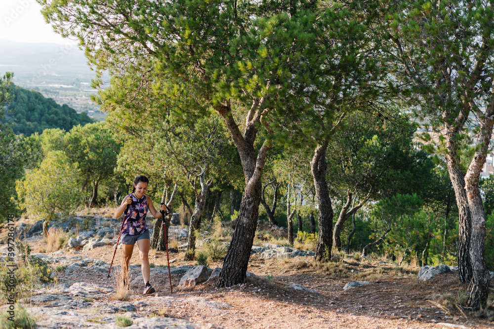 Active young female hiker with trekking poles climbing stony trail on mountain slope with green trees during training in nature