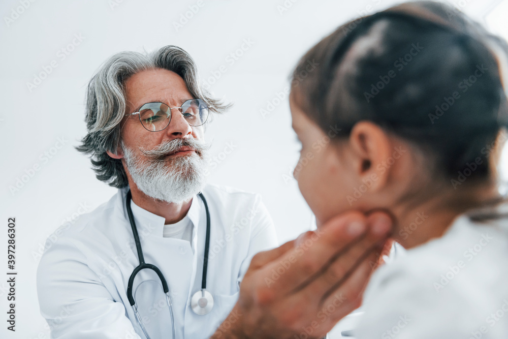 Little girl have a visit. Senior otorhinolaryngologist with grey hair and beard in white coat is indoors in clinic Little girl have a visit. Senior otorhinolaryngologist with grey hair and beard in white coat is indoors in clinic