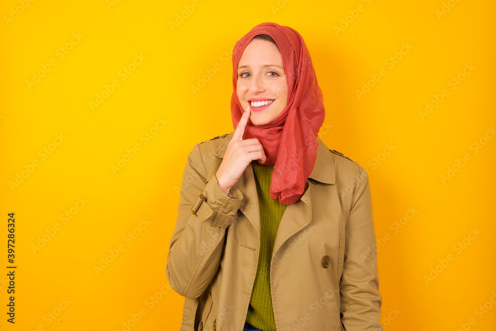 Carefree successful Young Caucasian Muslim woman wearing hijab standing against yellow wall touching jawline gazing camera tilting head grinning white teeth delighted. Dental care concept. Carefree successful Young Caucasian Muslim woman wearing hijab standing against yellow wall touching jawline gazing camera tilting head grinning white teeth delighted. Dental care concept.