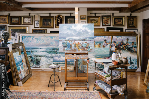 Interior of creative art workshop with painting on easel near shelf with tools and variety of ready framed pictures photo